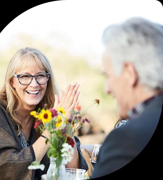 A woman in glasses laughing across a small table from a man over a bouquet of wildflowers, outdoors