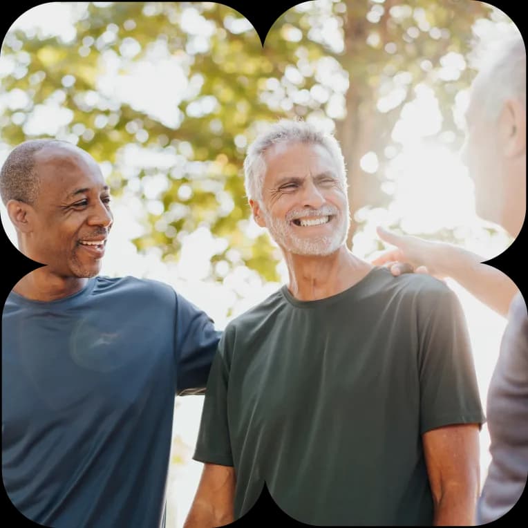 Three older men laughing together outdoors among sunlit green trees