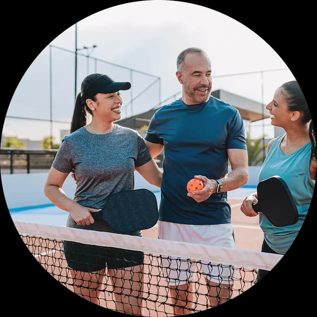 Three friends gathered at a pickleball net holding paddles and an orange ball